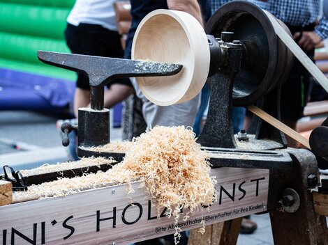 Eine Holzmaschine verarbeitet Holz zu einer Schüssel. Späne liegen auf dem Tisch und es sind Menschen im Hintergrund zu sehen.