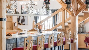 Eine gemütliche Bar in einem rustikalen Holzstil mit roten Barhockern. Im Hintergrund sind Holztreppen und dekorative Elemente zu sehen.