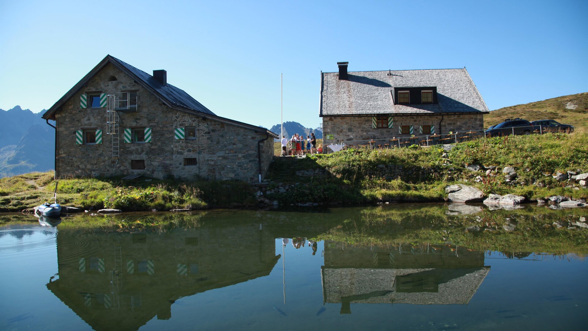 Eine malerische Berglandschaft mit zwei gemütlichen Häusern entlang eines ruhigen Sees. Die Gebäude spiegeln sich klar im Wasser und sind von grünen Wiesen umgeben.