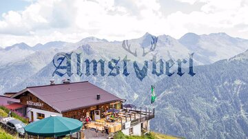 Ein Bergrestaurant mit einer großen Terrasse und Blick auf die umliegenden Berge. Die Landschaft ist grün und die Himmel ist bewölkt.