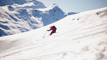 Ein Skifahrer fährt über eine schneebedeckte Piste in den Bergen. Der Himmel ist klar und die Berge sind mit Schnee bedeckt.