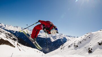 Ein Skifahrer springt über einen Schnee-Hügel in den Bergen. Der Himmel ist klar und die Sonne scheint strahlend.