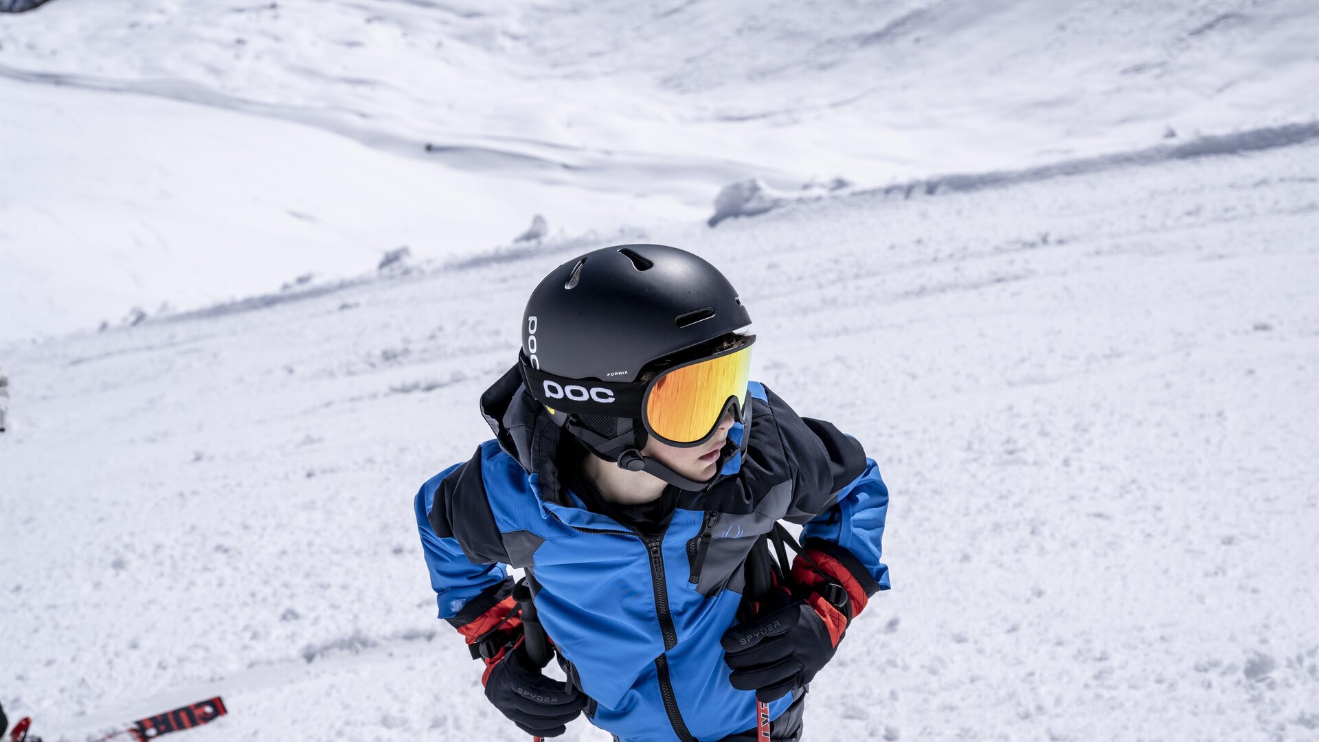A boy in a blue ski suit is wearing a helmet and ski goggles. He is standing on a snow-covered slope and looking up.