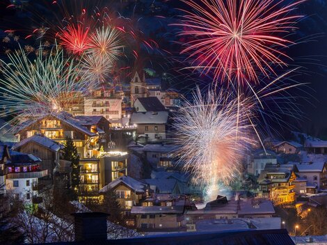 Eine festliche Szene mit bunten Feuerwerken über einem schneebedeckten Dorf. Die Häuser strahlen im Licht der Feuerwerke und schaffen eine festliche Atmosphäre.