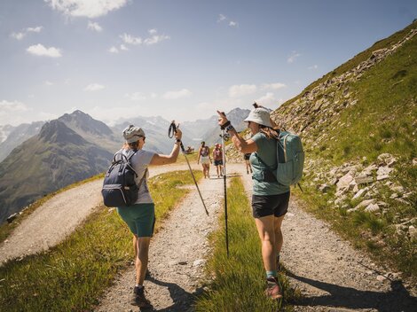 Zwei Wanderer auf einem Bergweg genießen die Natur. Im Hintergrund sind weitere Wanderer und Berge zu sehen.