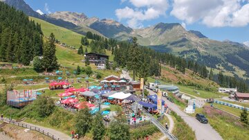 A lively mountain valley with many trees and a colorful terrace full of sunshades. In the background, majestic mountains and a bright blue sky can be seen.
