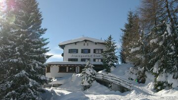 Ein malerisches Berghaus im Schnee, umgeben von hohen Tannenbäumen. Der klare Himmel und die Winterlandschaft verleihen der Szenerie eine einladende Atmosphäre.
