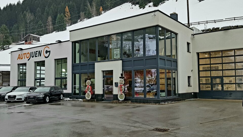 Ein modernes Autohaus mit großen Fenstern und einer modernen Architektur. Vor dem Gebäude stehen mehrere Fahrzeuge und im Hintergrund sind schneebedeckte Berge zu sehen.