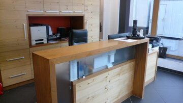 Ein moderner Empfangsbereich mit einem Holzschreibtisch und hellen Wänden. Im Hintergrund befinden sich Büromöbel und große Fenster, die Licht hereinlassen.