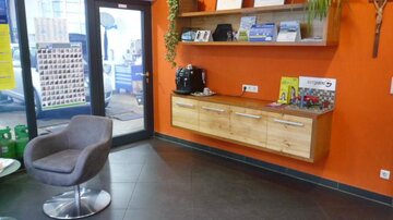 Ein moderner Empfangsbereich mit orangefarbener Wand und einer Holzregalwand. Im Raum befindet sich ein grauer Sessel und ein Kaffeeautomat.