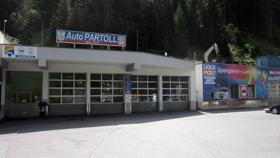 Eine Werkstatt namens Auto Partoll mit großen Garagentoren. Im Hintergrund sieht man Bäume und eine bunte Werbetafel.