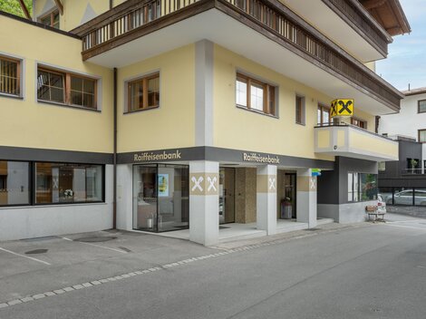 Ein modernes Bankgebäude mit gelber Fassade und großen Fenstern. Vor dem Gebäude befindet sich ein Parkplatz und eine Basketballanlage.