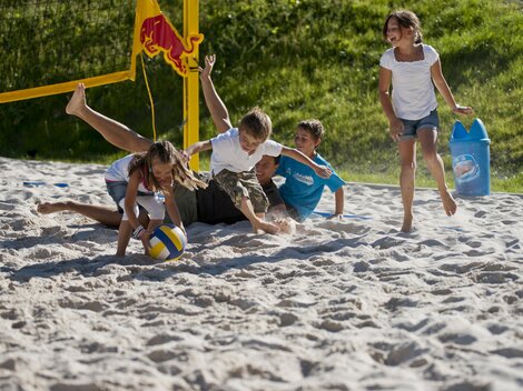 Eine Gruppe von Kindern spielt Beachvolleyball im Sand. Zwei Kinder lachen, während sie um den Ball kämpfen.