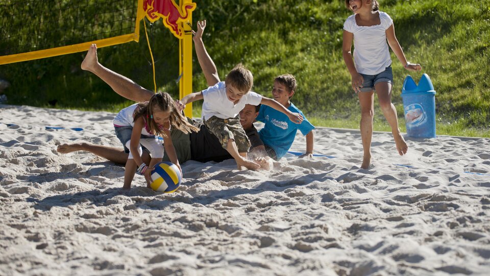 Eine Gruppe von Kindern spielt Beachvolleyball im Sand. Zwei Kinder lachen, während sie um den Ball kämpfen.