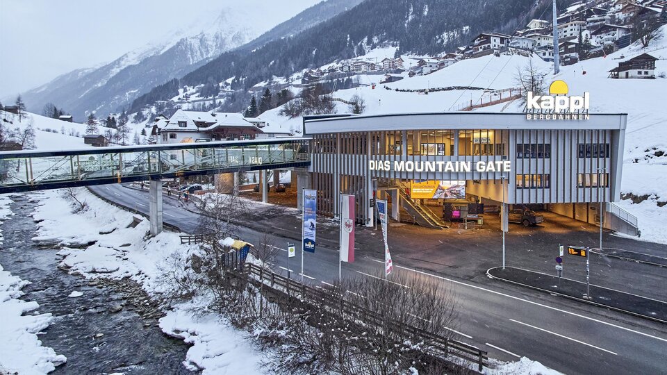 Ein modernes Gebäude mit der Aufschrift "DAS MOUNTAIN GATE" steht in einer schneebedeckten Landschaft. Im Hintergrund sind Berge und ein Fluss zu sehen.