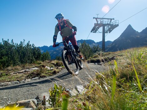 Ein Radfahrer fährt auf einem schmalen Weg in den Bergen. Im Hintergrund sind die Berggipfel und eine Seilbahn zu sehen.