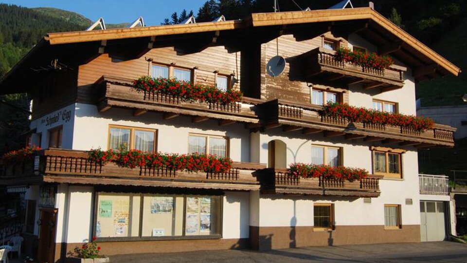 Ein traditionelles Holzgebäude mit vielen Balkonen, die mit roten Blumen geschmückt sind. Im Hintergrund sind grüne Berge zu sehen.