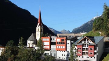 Ein malerisches Bergdorf mit einer Kirche und modernen Gebäuden. Die Umgebung ist von grünen Bäumen und hohen Bergen geprägt.