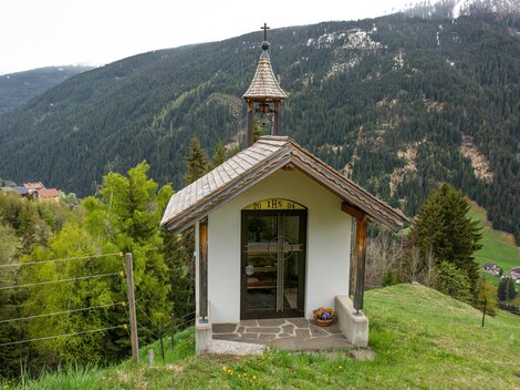 Eine kleine Kapelle steht auf einem Hügel, umgeben von Bäumen und Bergen. Im Hintergrund ist eine malerische Berglandschaft zu sehen. | © Bernhard Gruber – Photography