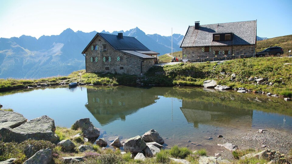 Eine malerische alpine Landschaft mit zwei Hütten am Ufer eines ruhigen Sees. Im Hintergrund erheben sich majestätische Berge unter einem klaren Himmel.
