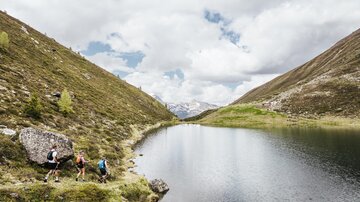 Eine ruhige Landschaft mit einem klaren See, umgeben von Bergen. Wanderer erkunden die grüne Umgebung unter einem bewölkten Himmel.