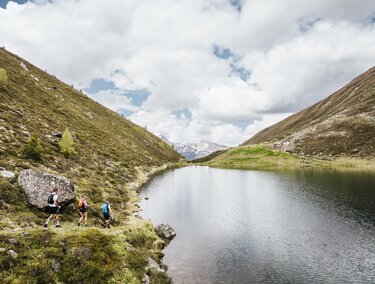 Eine ruhige Landschaft mit einem klaren See, umgeben von Bergen. Wanderer erkunden die grüne Umgebung unter einem bewölkten Himmel.