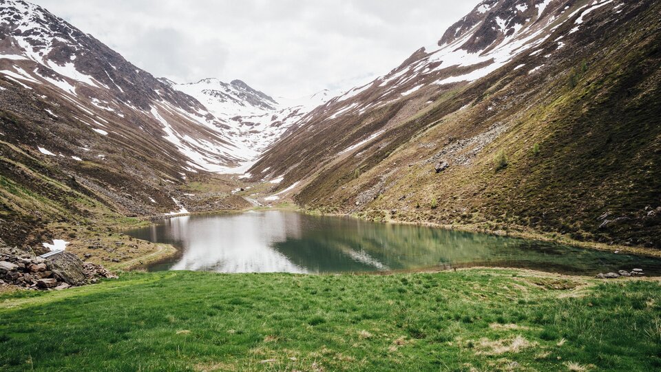 Ein ruhiger Bergsee umgeben von schneebedeckten Bergen. Die Landschaft ist grün und idyllisch.