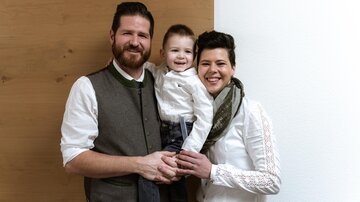 Eine Familie mit einem kleinen Kind steht zusammen vor einer hellen Wand. Alle tragen elegante, lässige Kleidung und lächeln freundlich.