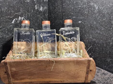 Drei Glasflaschen in einem Holzbehälter, umgeben von Heu. Die Flaschen sind mit Korken verschlossen und beschriftet.