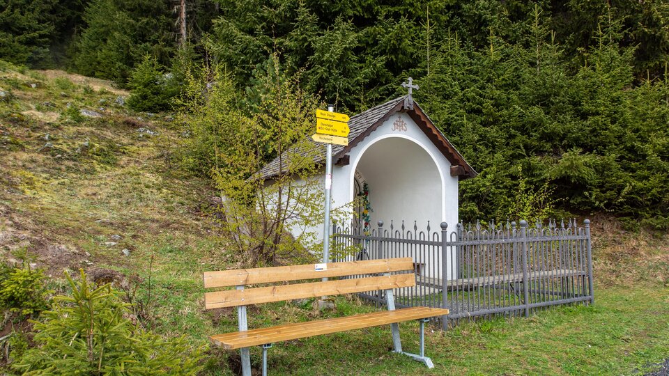 Eine kleine Kapelle steht in der Natur, umgeben von grünen Bäumen. Neben der Kapelle befindet sich eine Holzbank zum Sitzen und Ausruhen. | © Bernhard Gruber – Photography