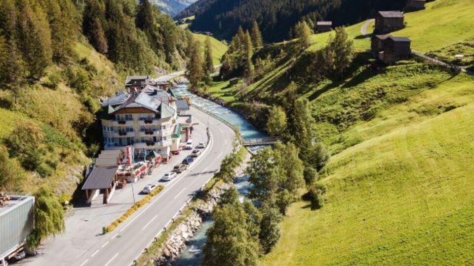 Eine malerische Landschaft mit einem Fluss, der neben einer Straße fließt. Im Hintergrund sind grüne Hügel und einige Chalets zu sehen.