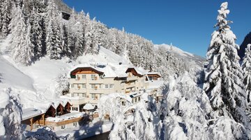 Ein malerisches Hotel in einer schneebedeckten Landschaft. Hohe Tannenbäume und blauer Himmel schaffen eine winterliche Atmosphäre.