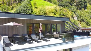 Eine moderne Terrasse mit Liegen und einem Sonnenschirm. Im Hintergrund sind grüne Hügel zu sehen.