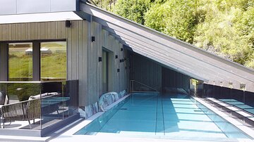 Ein modernes Haus mit einem stilvollen Pool und großen Fenstern. Die überdachte Terrasse bietet einen Blick auf die grüne Umgebung.