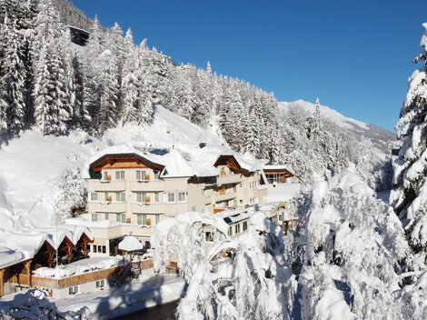 Ein malerisches Hotel in einer schneebedeckten Winterlandschaft. Hohe Tannenbäume und ein klarer blauer Himmel umgeben die Szene.