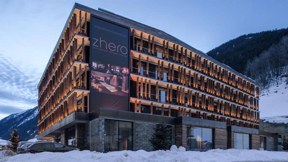 Ein modernes Hotel mit einer stilvollen Fassade und beleuchteten Balkonen. Umgeben von schneebedeckten Bergen und einem klaren Abendhimmel.