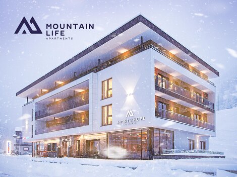 Ein modernes Apartmentgebäude im Schnee, umgeben von einer winterlichen Landschaft. Die Fassade ist in hellen Tönen gehalten und verfügt über große Fenster.
