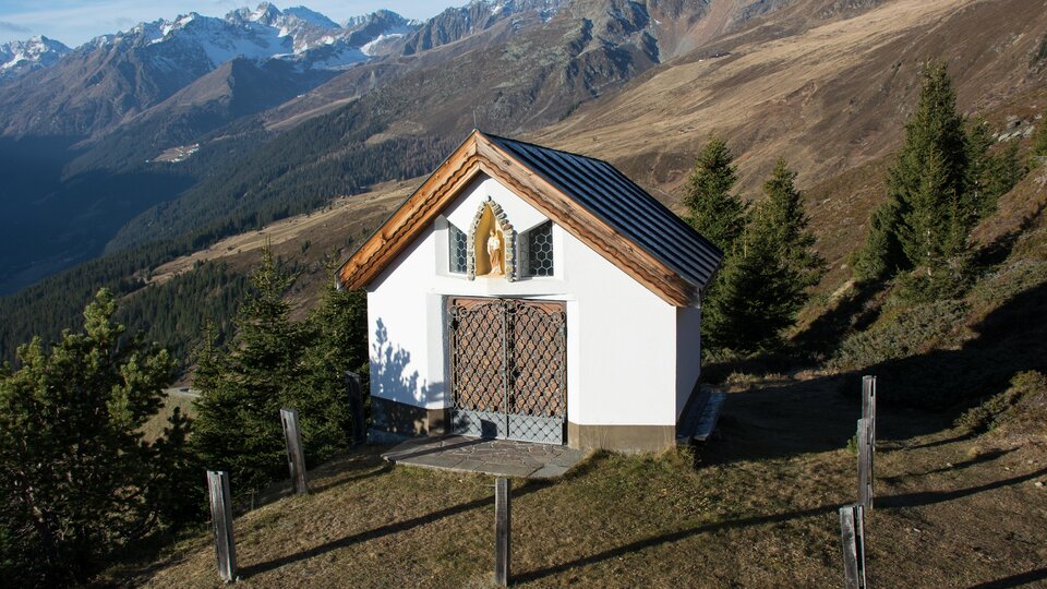Eine kleine Kapelle auf einem Hügel, umgeben von Bergen und Wäldern. Die Landschaft zeigt klare Himmel und Berggipfel im Hintergrund. | © Bernhard Gruber – Photography