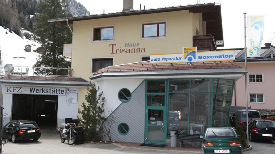 Ein gemütliches Gebäude mit der Aufschrift "Haus Trisanna" und einer Werkstatt für Auto-Reparaturen. Im Vordergrund sind einige Autos und Fahrräder zu sehen.