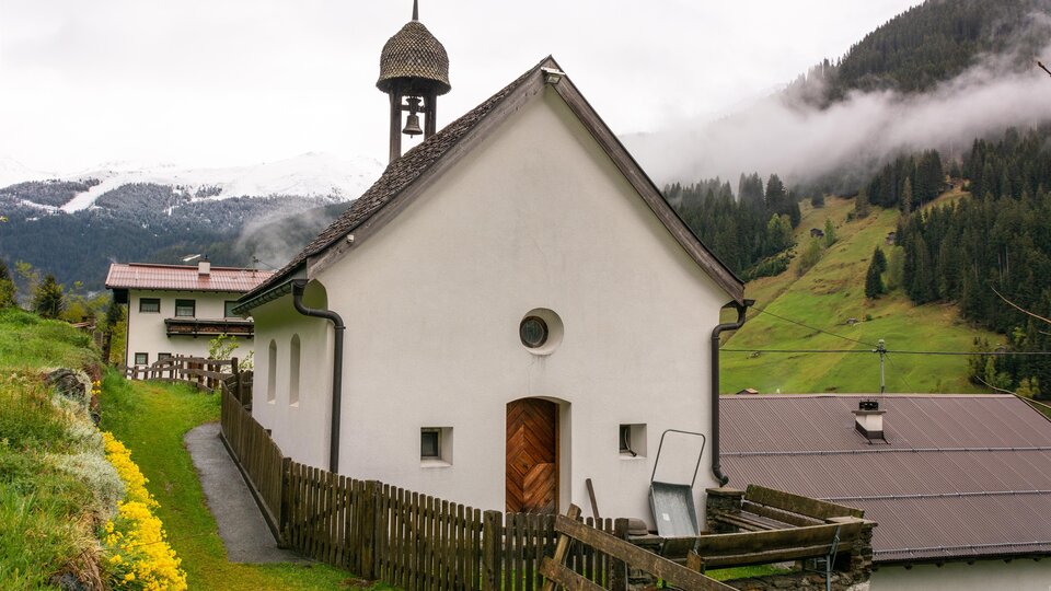 Eine kleine, weiße Kirche mit einem Glockenturm steht in einer malerischen Landschaft. Im Hintergrund sind grüne Hügel und bewölkter Himmel sichtbar. | © Bernhard Gruber – Photography