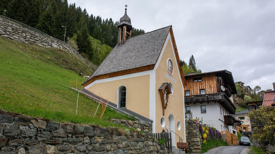 Eine kleine, orangefarbene Kapelle steht am Hang, umgeben von Grün. Im Hintergrund sind typische alpine Holzhäuser zu sehen. | © Bernhard Gruber – Photography