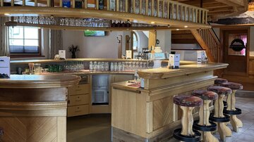 Eine gemütliche Bar aus Holz mit mehreren Hockern und einem gut gefüllten Regal. Das Ambiente ist warm und einladend.