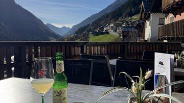 Ein schöner Tisch auf einer Terrasse mit Blick auf die Berge. Auf dem Tisch stehen ein Glas mit Getränk, eine Flasche, eine Tasse Kaffee und eine kleine Pflanze.