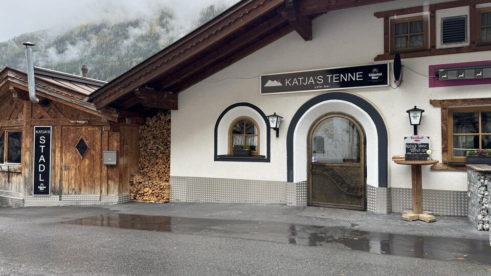 Ein traditionelles alpenländisches Gebäude mit einem einladenden Eingang. Neben dem Gebäude befindet sich ein Holzstapel und ein Schild mit der Aufschrift "STADL".