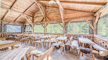Ein gemütlicher Innenraum eines Restaurants mit Holzstühlen und -tischen. Große Fenster bieten einen Blick auf die Natur.