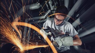 Ein Handwerker arbeitet mit einer Schleifmaschine und erzeugt Funken. Er trägt Schutzbrille und Arbeitshandschuhe in einer industriellen Umgebung.