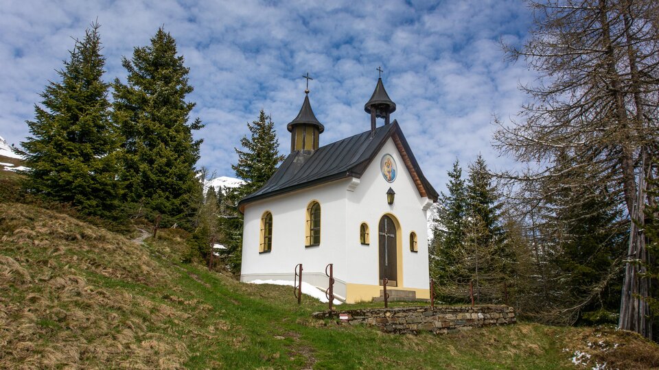 Eine kleine, charmante Kapelle steht auf einem Hügel, umgeben von grünen Wiesen und hohen Tannen. Der Himmel ist klar und zeigt einige Wolken. | © Bernhard Gruber – Photography