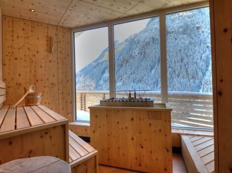 Ein gemütlicher Wellnessbereich aus Holz mit Blick auf schneebedeckte Berge. Das Licht strömt durch große Fenster und schafft eine entspannende Atmosphäre.