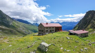 Eine Berghütte umgeben von grünen Wiesen und majestätischen Bergen. Der Himmel ist klar mit einigen Wolken.