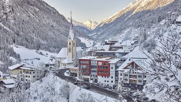 Ein malerisches Winterdorf mit schneebedeckten Bergen und einer Kirche im Vordergrund. Die Straßen sind ruhig, umgeben von weißen Tannen und charmanten Häusern.